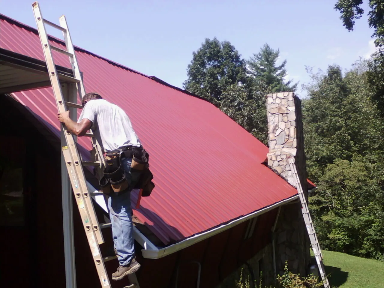 Roofer on a ladder beside a red metal roof and stone chimney — Soffit & Fascia Repair by Roofing Brothers in Fort Loudon